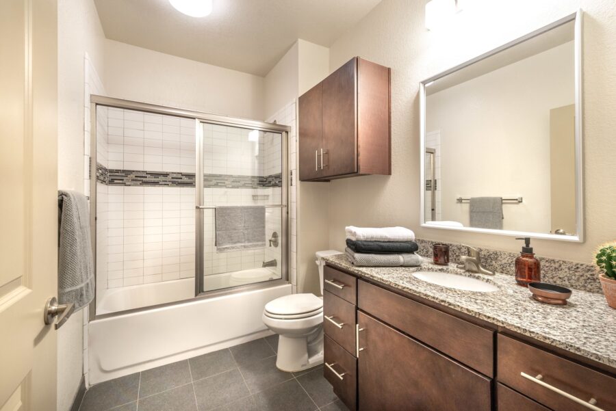 Interior of student apartment bathroom with glass wall around tub and wooden cabinetry